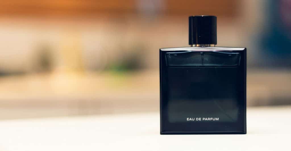 A dark, rectangular perfume bottle with a black cap stands on a light surface with a blurred, warm-toned background.