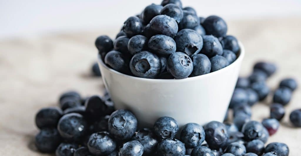 A white bowl overflowing with fresh blueberries, with more blueberries scattered around it on a light-colored cloth.