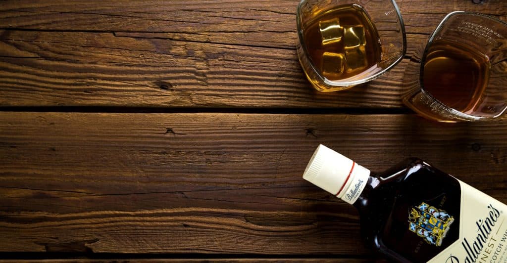 A bottle of Ballantine's Finest Blended Scotch Whisky lies on a dark wooden table next to two glasses, one containing ice and amber liquid.