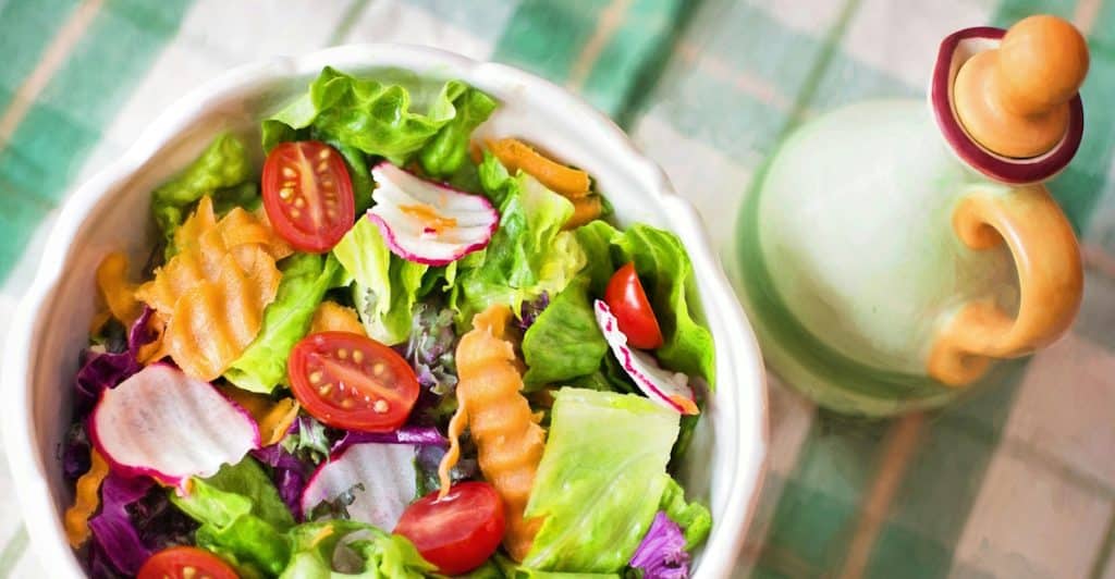 A white bowl filled with a colorful fresh green salad next to a green and orange dressing cruet, all on a green and white plaid tablecloth.