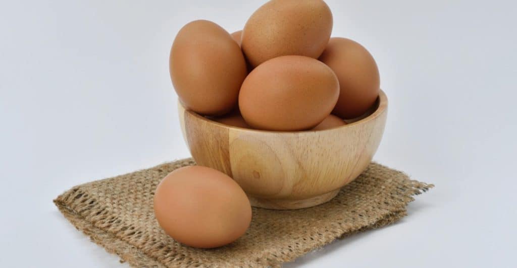 Several brown eggs are stacked in a rustic wooden bowl, which rests on a piece of woven beige textile, with one egg placed beside the bowl on a white surface.
