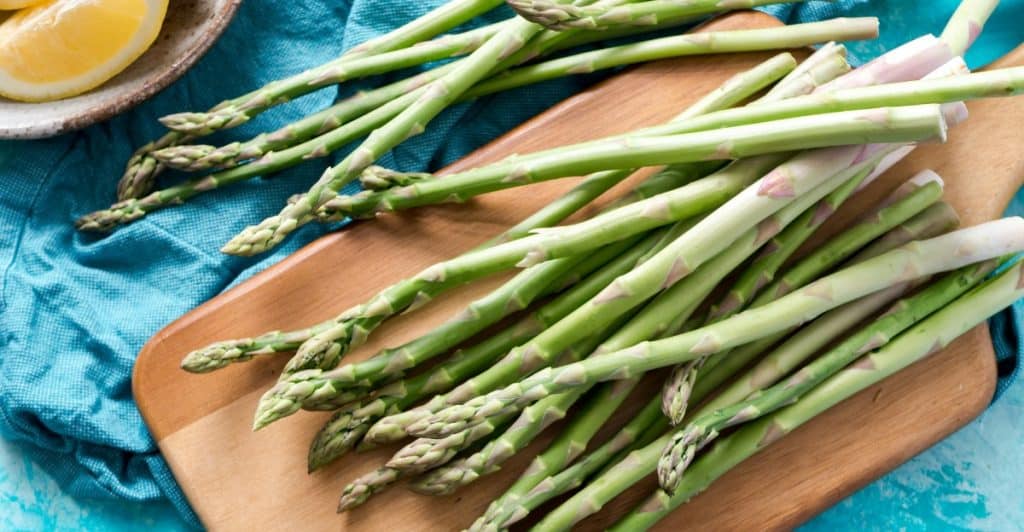 A bunch of fresh green asparagus spears arranged on a wooden chopping board with lemon slices nearby.