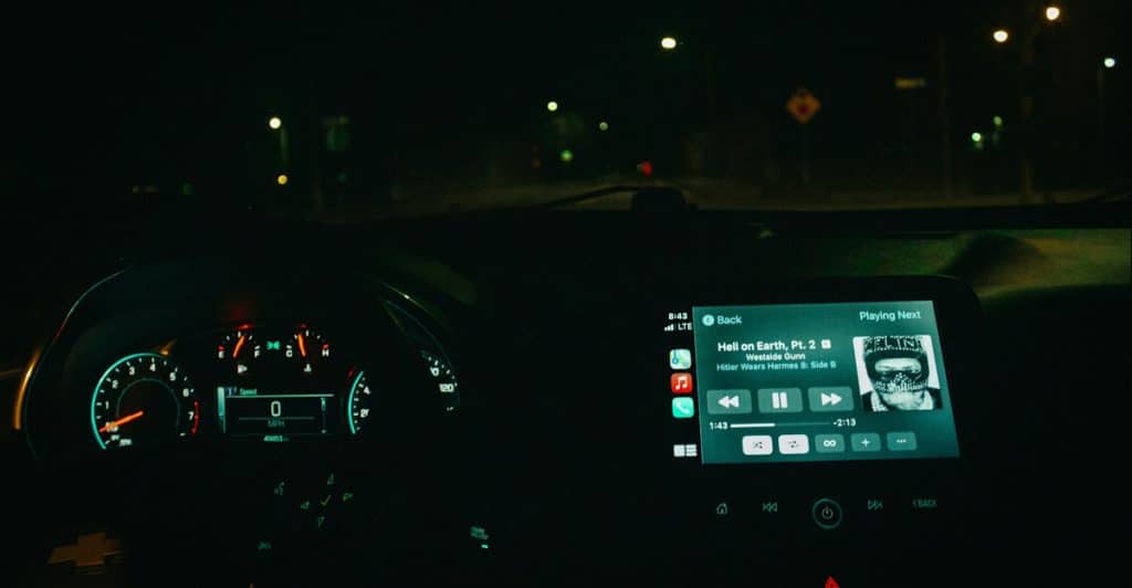 The interior of a car at night showing the dashboard with illuminated gauges and a central display screen playing music.