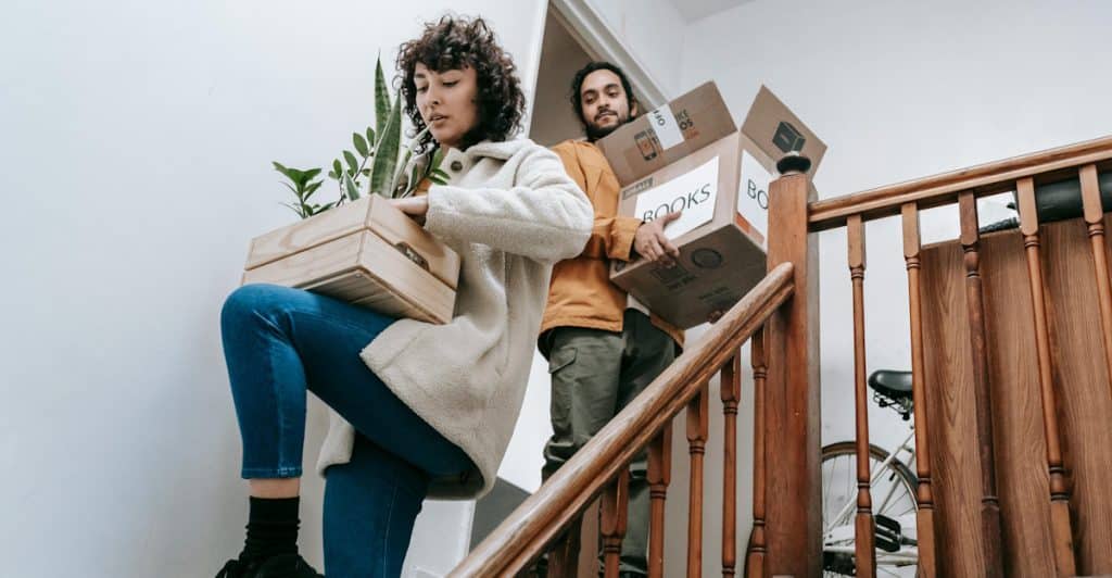 A woman and a man carrying cardboard boxes, one labeled "BOOKS," as they walk down a wooden staircase inside a house.