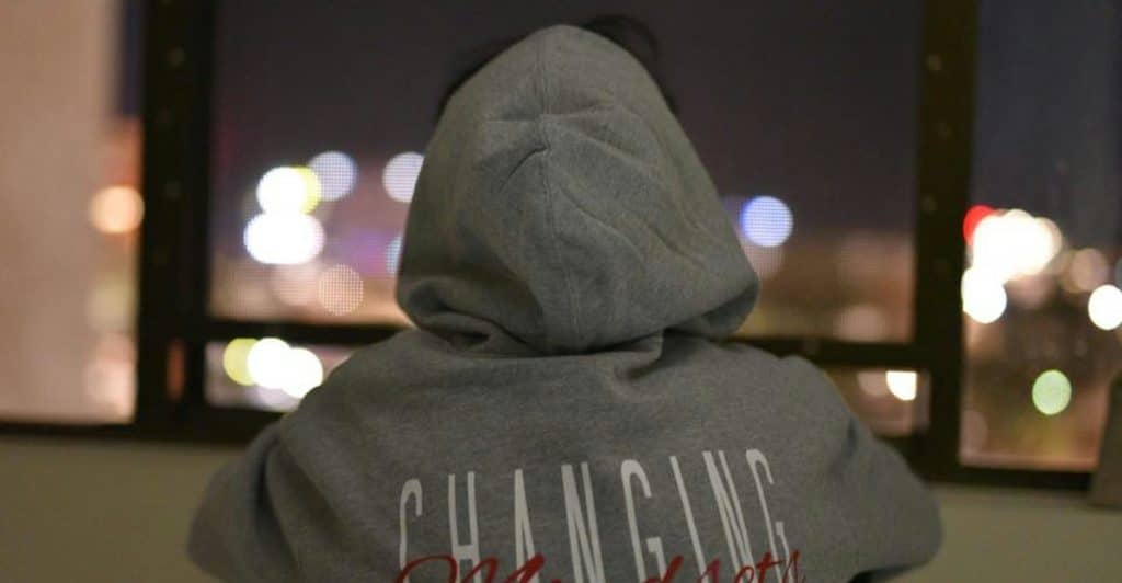The back of a person wearing a gray hoodie with "CHANGING Mindset" written on it, looking out a window at a blurry cityscape at night.