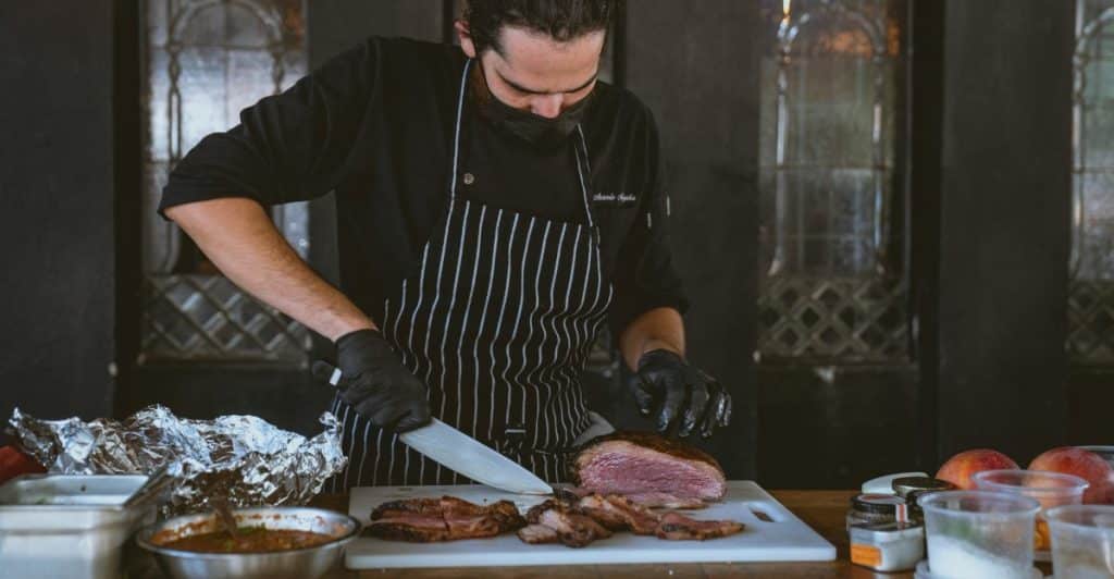 A chef wearing black gloves and an apron, meticulously carving slices from a large piece of roasted meat on a white cutting board, with various dishes around him.