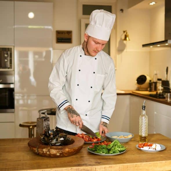 A man in a chef's hat and white uniform chopping vegetables on a wooden counter in a kitchen.