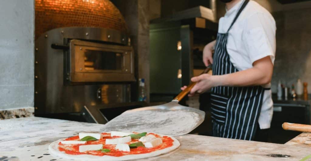 A chef wearing a black mask and a striped apron standing in front of a large brick pizza oven, preparing a pizza with fresh mozzarella and basil on a wooden peel.