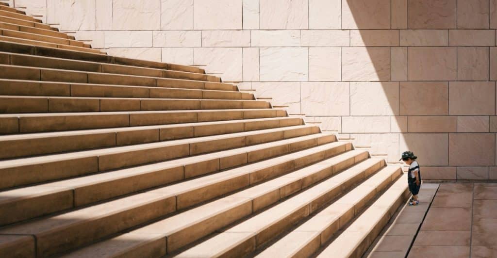 A small child in a cap and backpack looking up a very long, wide stone staircase.