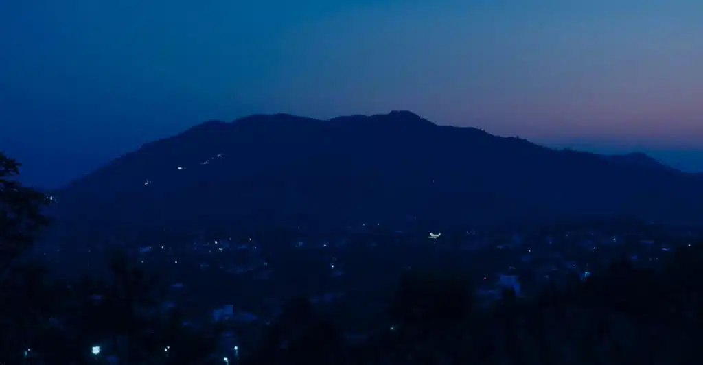 A dark silhouette of mountains against a deep blue sky overlooking a city with scattered lights at dusk.