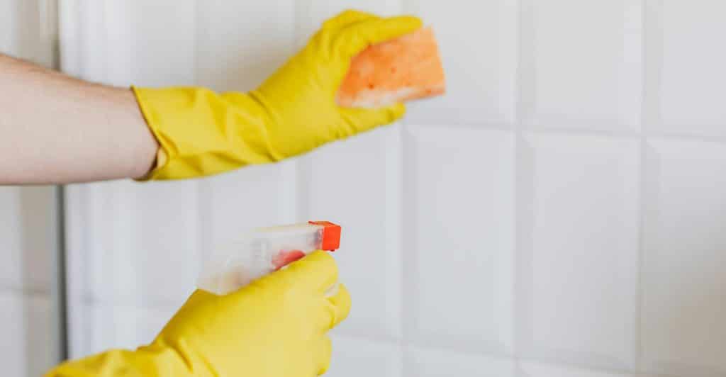 Hands in yellow gloves holding an orange sponge and a spray bottle, cleaning white tiled walls in a bathroom.