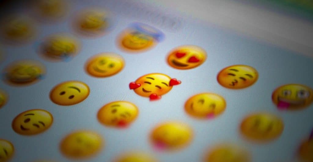 A close-up, slightly blurred image of a digital screen displays various yellow emojis, with one prominent heart-eyed emoji in the center, surrounded by others like a winking face and a kissing face.