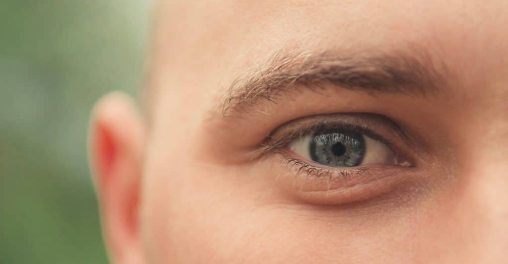 A close-up of the right side of a man's face, focusing on his blue eye and ear, with a blurred green background.