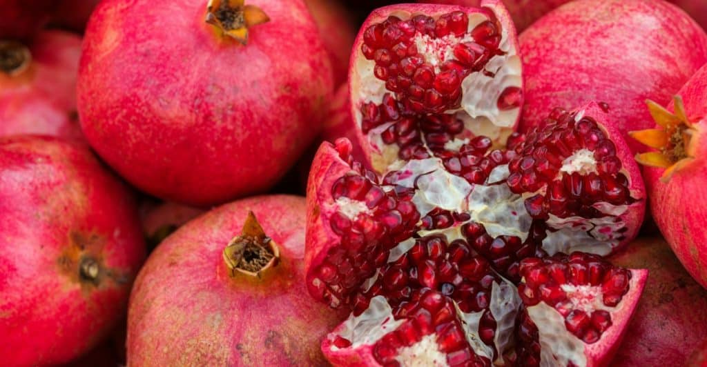 A close-up picture of many red pomegranates, with one fruit broken open to show its seeds.