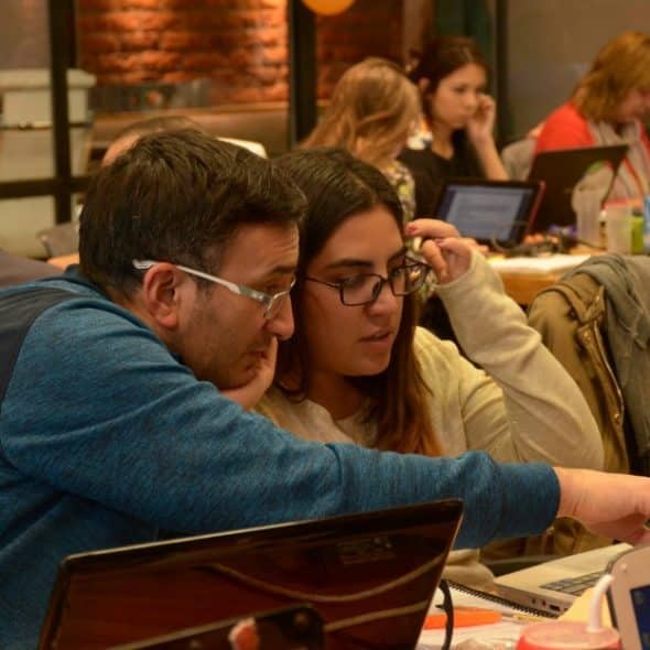 A group of people working on laptops in a collaborative coding workshop, with one man pointing at a screen while assisting a woman.