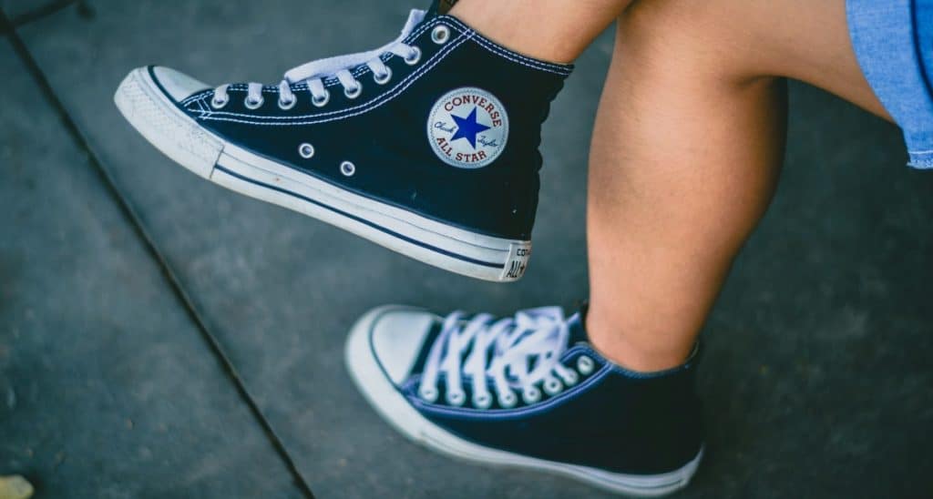 A person wearing black Converse Chuck Taylor All-Star high-top sneakers with white laces, sitting with their feet suspended.