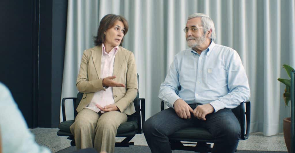 A woman in a tan blazer gesturing with her hand while talking to a man sitting beside her, with a laptop in front of them and a striped curtain background.