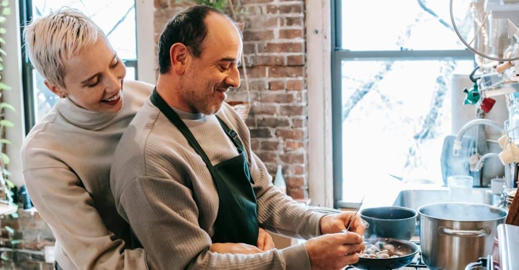A woman with short blonde hair hugging a man from behind, both smiling as he stirs food in a pan on a stove in a bright kitchen.
