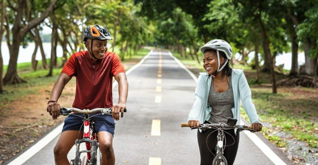 A joyful couple, both wearing helmets, ride bicycles side-by-side on a paved path lined with tall green trees, smiling and looking at each other, with a body of water visible in the background.