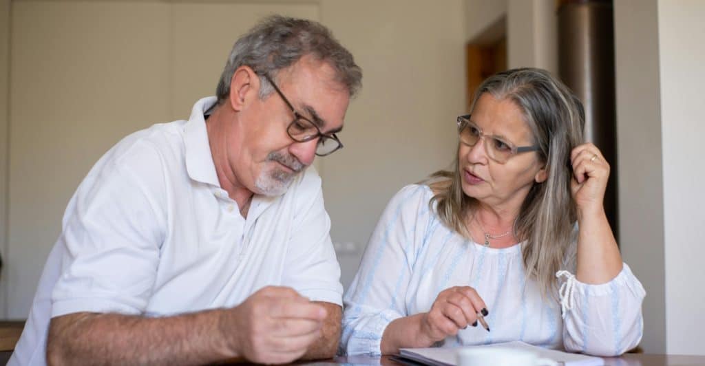 An older couple, both wearing eyeglasses, sitting at a table with papers and a pen, appearing to be seriously discussing and completing documents.