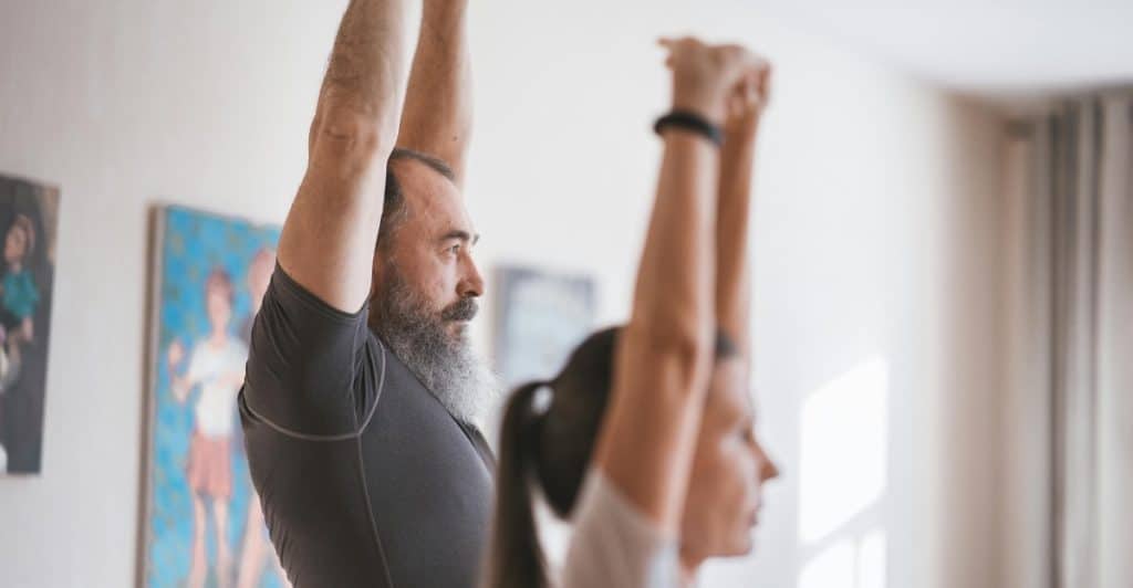 A man with a long beard and a woman, both wearing athletic wear, are shown from the side in a home setting, with their arms raised above their heads, engaged in a stretching exercise.