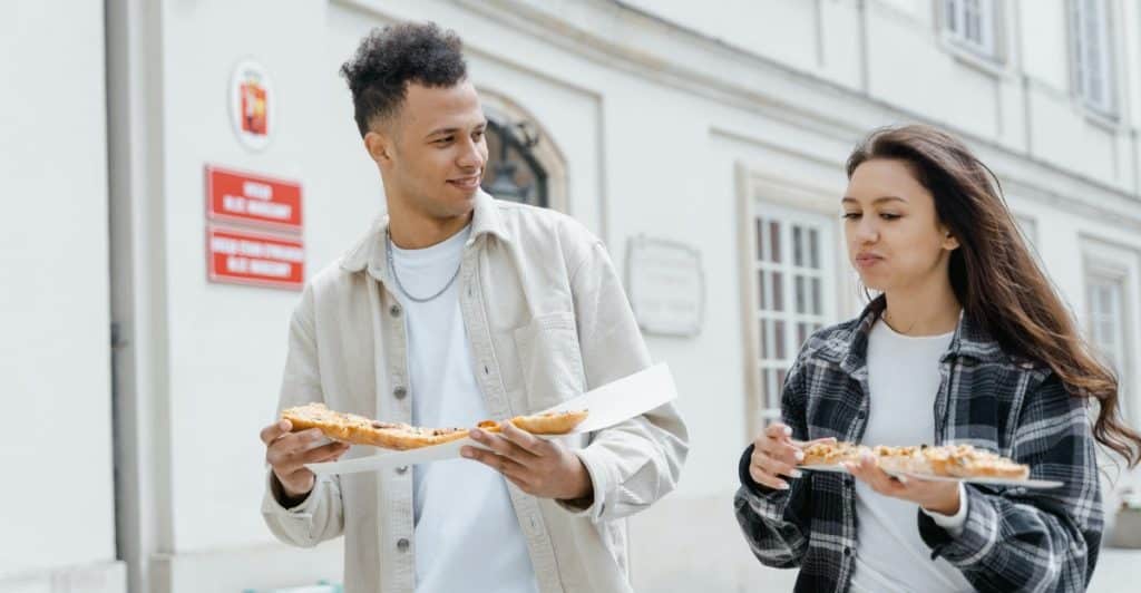 A man and a woman eating pizza slices outdoors in a city setting.