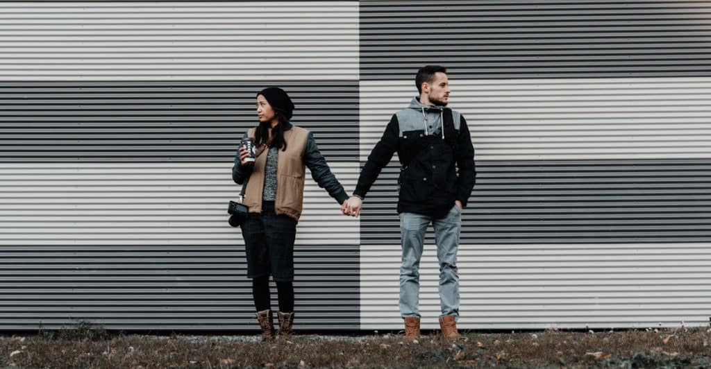 A couple standing outdoors, holding hands and looking away from each other, in front of a striped black and white wall.