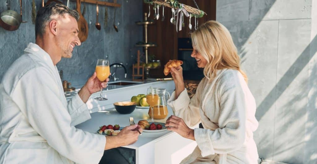 A man and a woman in white robes sitting at a kitchen counter, having breakfast and smiling at each other.