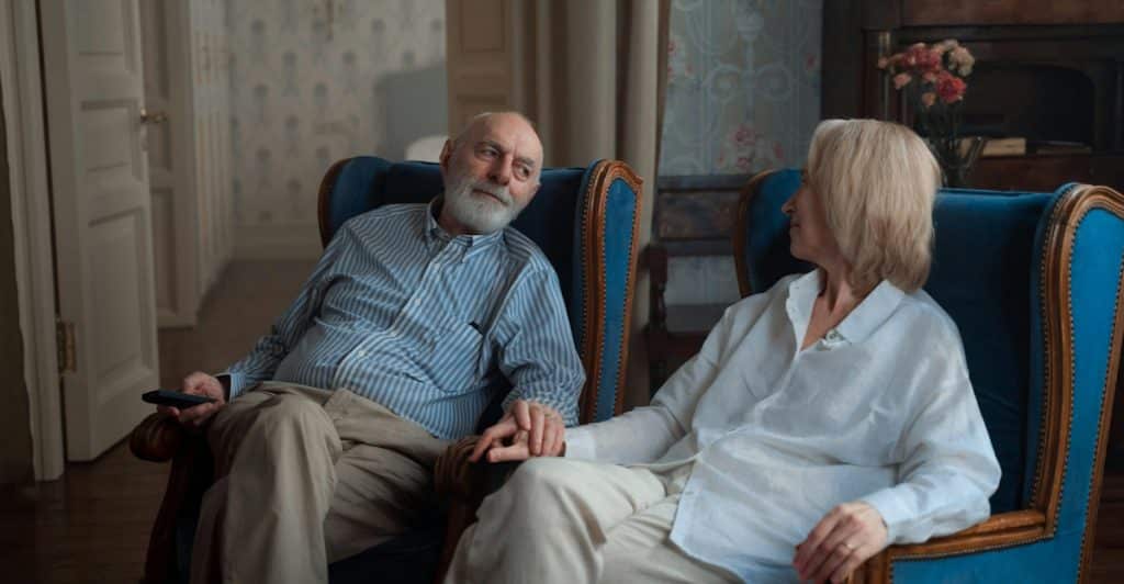 An older couple sitting in separate blue armchairs in a warmly lit living room, holding hands and looking at each other affectionately.