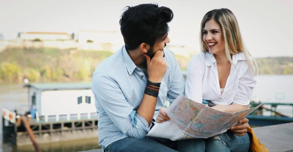 A man and a woman sitting outdoors, both looking at each other, with the woman smiling and holding a map.
