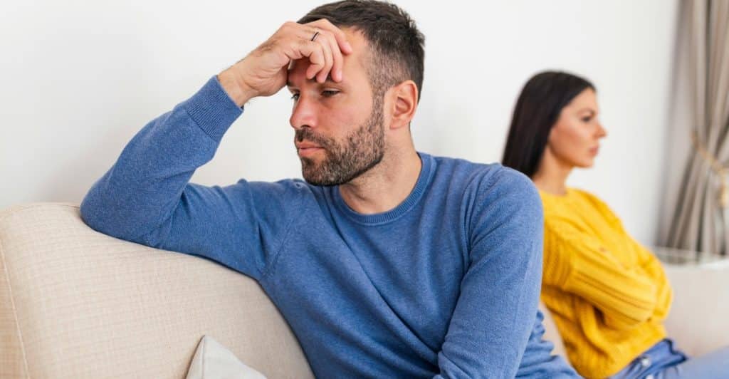 A man with a beard sitting on a couch with his hand on his head, and a woman sitting with her back to him, both appearing to be in a disagreement