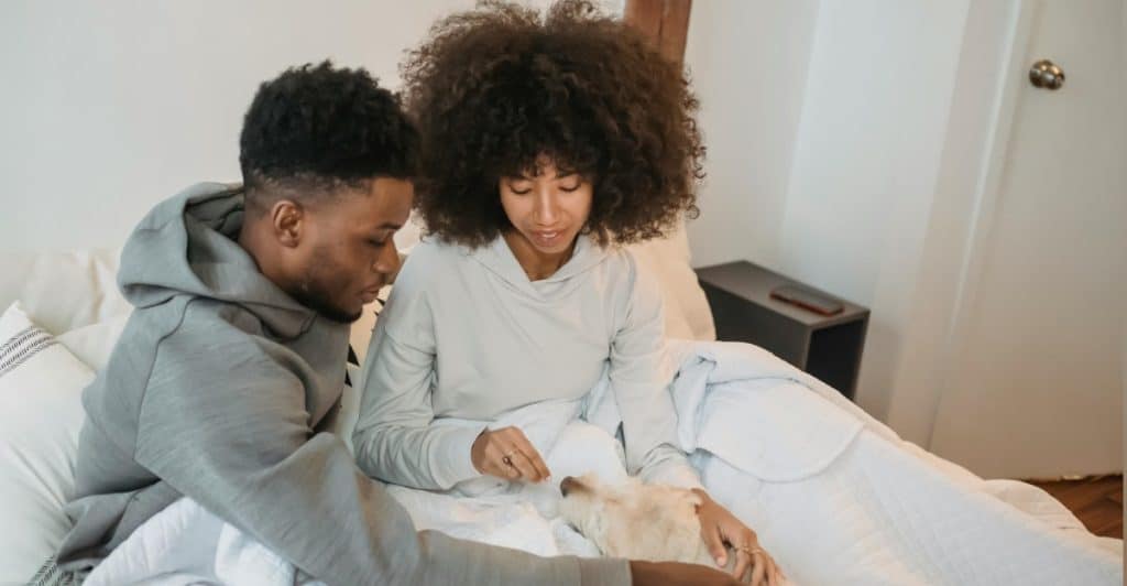 A couple petting a dog while sitting in bed.
