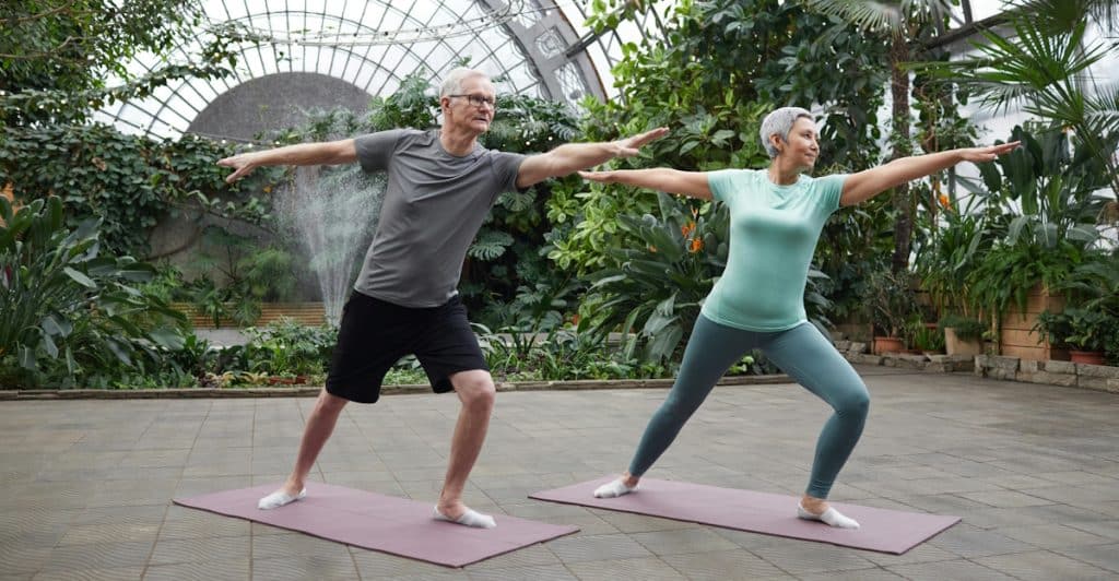 An older couple, a man and a woman, standing on yoga mats in a spacious indoor botanical garden with glass ceilings, practicing a yoga pose with outstretched arms.