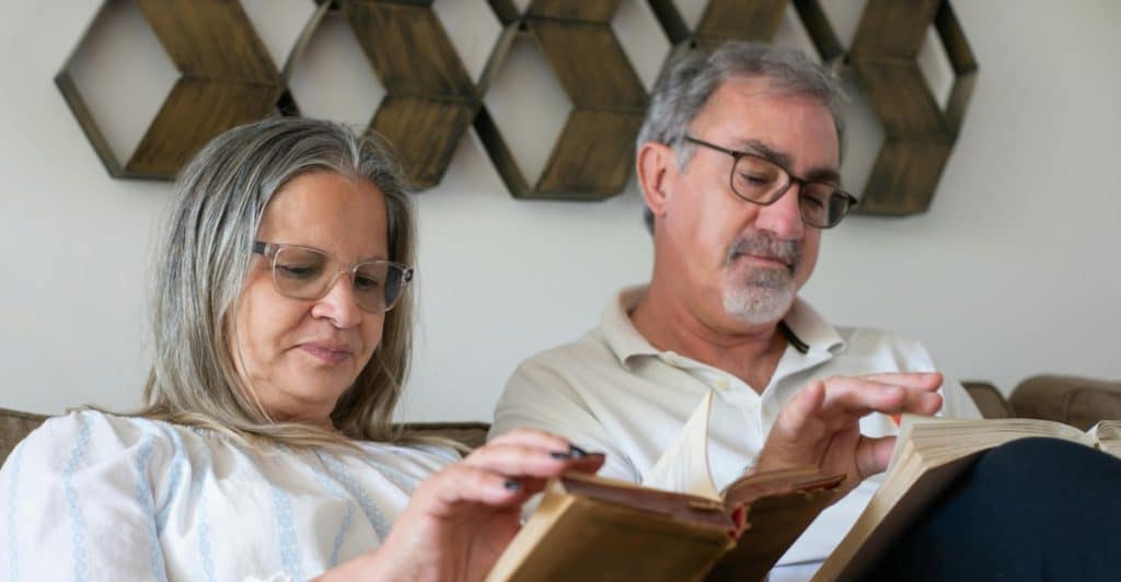 An older couple, both wearing eyeglasses, sitting closely together on a sofa and deeply engrossed in reading their individual books.