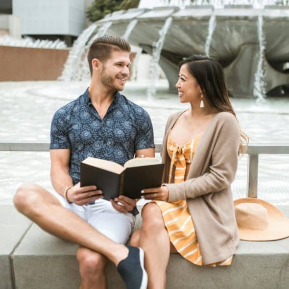 A man and woman sitting outside by a fountain reading a book together.