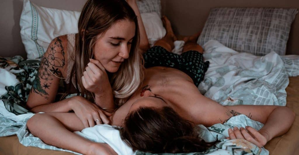 A woman with tattoos on her arm lying on her stomach in bed, looking at a man who is lying on his back.