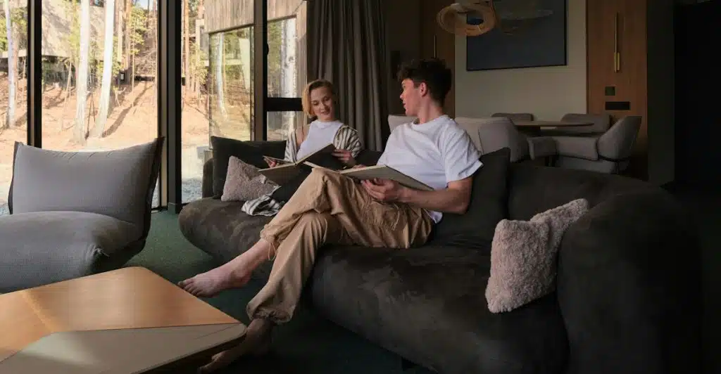 A man and a woman sitting comfortably on a dark gray sofa in a modern living room, both holding books, enjoying a moment of rest.