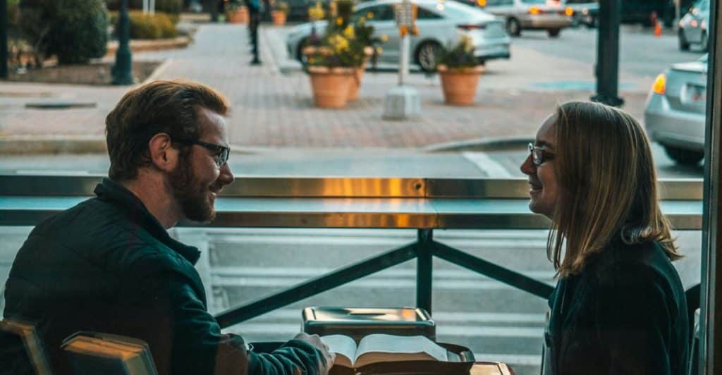 A man and a woman sitting at an outdoor table with a book, looking at each other and smiling, with a street and buildings in the background.
