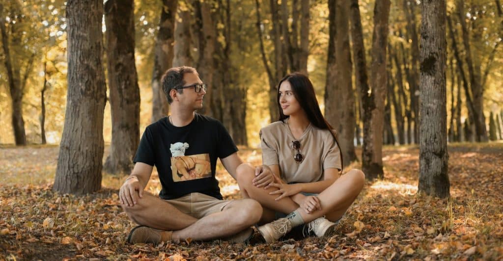 A couple is seated cross-legged on the ground amidst fallen autumn leaves in a park, facing each other and smiling, surrounded by tall trees with golden foliage.