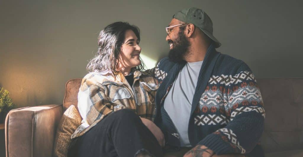 A man wearing a cap and glasses and a woman with dark hair sitting on a couch, looking at each other and smiling.