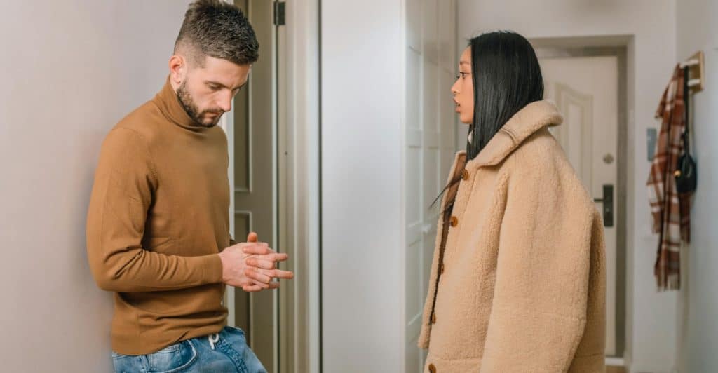 A man leaning against a wall and a woman standing in a hallway, both looking at each other.