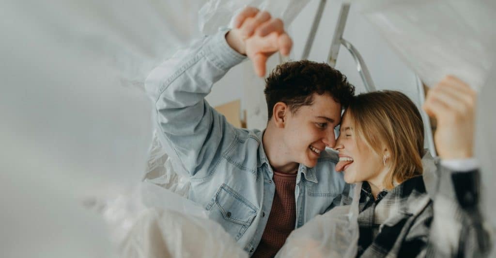 A cheerful couple looking at each other and smiling from underneath clear plastic sheeting, suggesting a playful or intimate moment.