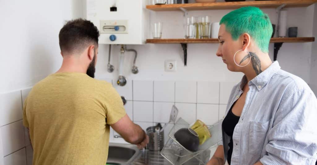 A man with a beard washing dishes at a kitchen sink, while a woman with short green hair stands beside him, holding a white towel.