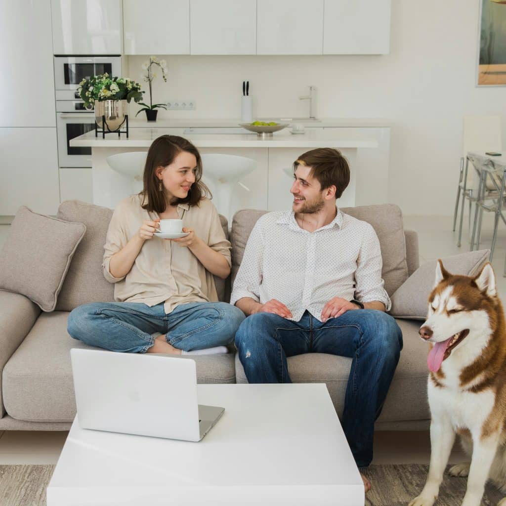 A man and a woman are sitting on a sofa in a living room with a husky dog sitting beside them.