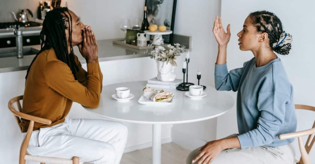 A man and woman sitting across from each other at a round table in a kitchen having a heated discussion with food and coffee on the table