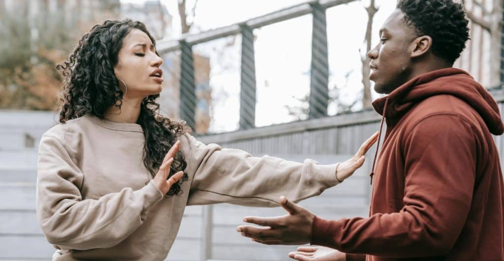A man and a woman arguing outdoors, with the woman gesturing with her hands.
