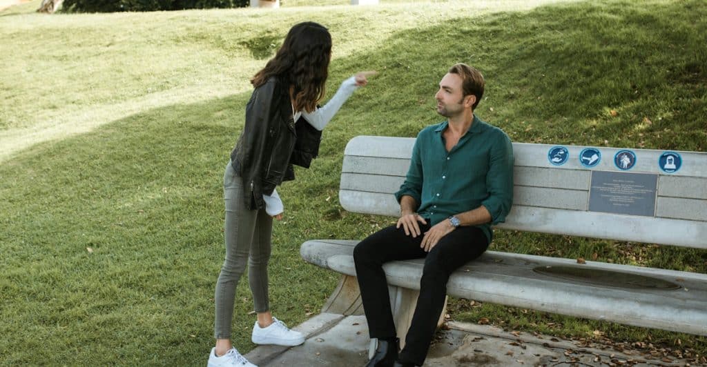 A woman pointing at a man who is sitting on a park bench.