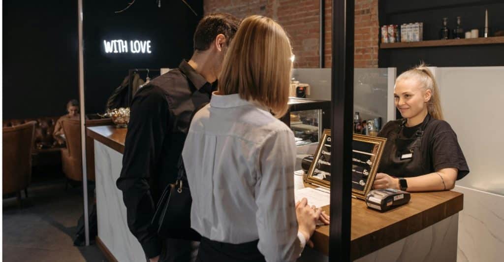 A man and a woman standing at a counter in a cafe, looking at a menu, with a smiling barista behind the counter.