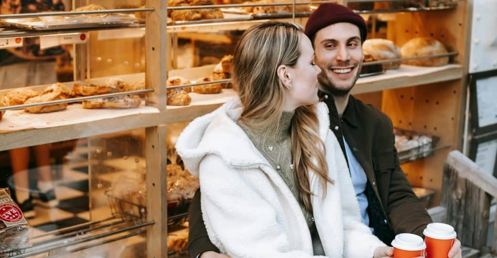 A smiling couple sitting at an outdoor table in front of a bakery, holding coffee cups, with the man's arm around the woman.