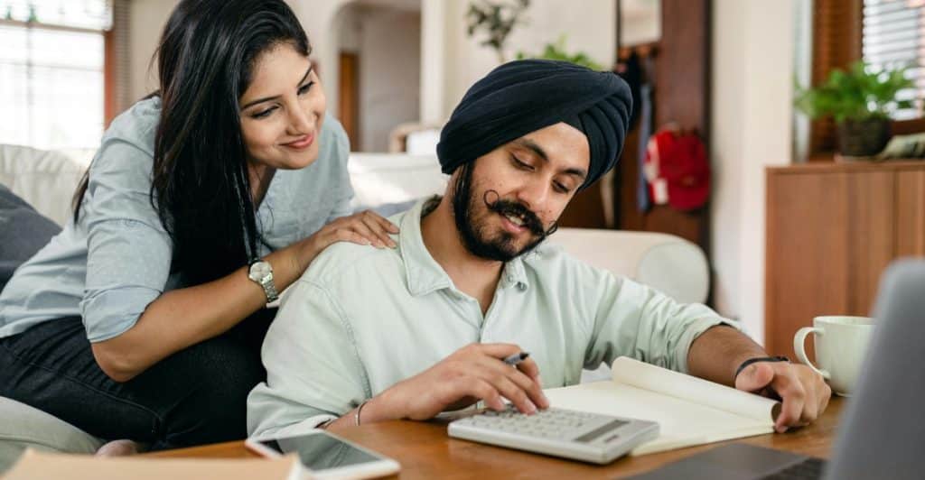 A man and a woman sitting at a table, with the man looking at a notebook and using a calculator, and the woman leaning on his shoulder and smiling.